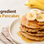 A stack of golden-brown, thin banana pancakes plated on a ceramic dish, topped with fresh strawberries and a side of Greek yogurt.